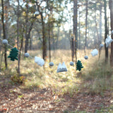 The wool felt mountain and cloud garland is suspended, set against a sunlit forest backdrop, evoking a sense of nature. This 3D-designed garland features alternating green felt trees, gray and white felt mountains, and soft felt clouds. Each element is separated by small gray felt balls, creating a charming display.