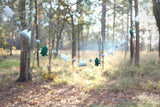 A wool felt mountain and cloud garland is strung across a blurred forest background, evoking a calm and playful atmosphere. It features alternating white clouds, grey mountains with white snow caps, and dark green fir trees. Small felt balls separate these delightful elements along the white string.