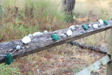 A handmade wool felt Mountain & Cloud Garland is draped across a weathered wooden fence, set against a natural, wooded backdrop, creating a rustic and whimsical atmosphere. This charming 3D-designed felt garland features alternating white felt clouds, grey felt balls, and stylized green felt trees on a thin white string.