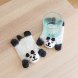 Two handcrafted felt panda coasters, part of a set, are displayed on a light wooden surface, one holding a clear glass of water. These unique wool felt coasters are shaped like pandas, featuring white bodies with black ears, eyes, and feet. The needled felted facial details enhance their charm, reflecting the ethical craftsmanship of Nepalese female artisans using 100% sheep wool.