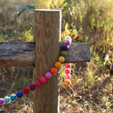 A vibrant wool felt rainbow pompom ball garland is draped over a weathered gray wooden fence post, with bright sunlight filtering through tall grasses in the background. The approximately 4-foot long garland features soft, 3D-designed wool felt balls in a rainbow spectrum of pink, orange, yellow, green, purple, blue, and light blue.