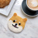 A cheerful wool felt Shiba Inu dog coaster rests on a smooth marble surface, next to a coffee cup and croissant. This handmade coaster, crafted from 100% sheep wool, features tan and white fur with needle-felted black eyes, a nose, and a pink tongue, capturing the dog's joyful expression. Approximately 4 inches in size, this unique coaster is ethically handmade by Nepalese female artisans.