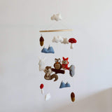 A wool felt woodland animal baby mobile is displayed against a plain white background. It features various needle-felted 3D figures crafted from 100% New Zealand wool, including white clouds, blue mountains, brown acorns, and woodland creatures like a squirrel, owl, fox, and rabbit. The mobile uses a soft, muted color palette, creating a gentle, comforting, and whimsical atmosphere perfect for a nursery.