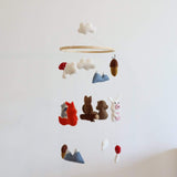 This charming wool felt woodland animal baby mobile hangs against a plain white wall. It features soft, needle-felted shapes of white clouds, blue mountains, brown acorns, and various woodland creatures like a red fox, brown bears, a gray squirrel, and a white rabbit. Suspended by thin white strings, the design uses muted, earthy colors, creating a calm and playful atmosphere.