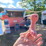 A needle felted flamingo finger puppet is gently held in a female hand at a whimsical outdoor market. This handmade felt finger puppet features a soft pink body and a distinctive black and white beak. The background shows a charming pink and white vendor booth adorned with colorful balloons under a sunny sky, creating a festive atmosphere.