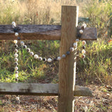 A Gray Felt Ball Garland is draped over a weathered wooden fence, set in a field of tall grass, creating a rustic outdoor scene. This felt ball garland features wool felted balls in varying shades of gray, cream, and brown, evenly spaced on a thin white string. The visible texture of the wool felt adds a soft, handcrafted feel.