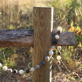 A gray wool felt ball garland is casually draped over a weathered wooden fence post, set against a sunlit, grassy field. This rustic garland features evenly spaced, spherical wool felt balls in varying shades of gray, brown, and white, adding a warm touch to the scene.