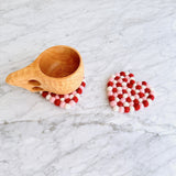 A set of four heart-shaped wool felt Love coasters, crafted from red, white, and pink organic lamb wool felt balls, rests on a bright marble surface. One handcrafted felt coaster supports a light wooden mug, while another displays its charming design. These unique, ethically made coasters reflect warmth and artisanal quality.