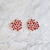 Two heart-shaped Love coasters rest on a white marble surface with gray veins. These handmade felt coasters are crafted from small, alternating red, pink, and white organic wool felt balls arranged in a grid pattern. Ethically made by Nepalese female artisans, these durable and charming coasters add a warm, romantic touch to any home decor.