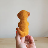 A handmade needle felted meerkat finger puppet, crafted from soft gold and orange felt, is held up by a female hand against a bright, neutral background. This unique felt toy is seen from the back, showcasing the meerkat's small, rounded ears and bushy tail. This fair-trade finger puppet for kids embodies artisan craftsmanship.
