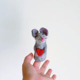 A charming gray mouse finger puppet holding a red heart is held by a female hand against a plain white backdrop. This unique needle-felted wool felt mouse finger puppet, handmade in Nepal, features a bright pink nose, inner ears, and whiskers. It's an endearing and detailed felt toy for kids.