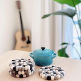 Neutral gray felt ball coasters are displayed on a light wooden table, creating a cozy and natural setting. This set of four round, multi-toned gray wool felt coasters is ethically handmade from organic lamb wool. Their tactile texture comes from numerous hand-stitched felt balls in varying shades of gray, white, and black. One coaster rests flat, with three more stacked beside it.