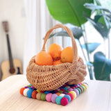 The Rainbow Square Felt Ball Pot Trivet rests on a light-colored wooden surface, holding a small, light-brown wicker basket filled with vibrant oranges. This handmade pot trivet, crafted from 100% organic lamb wool, features rows of hand-stitched felt balls in a spectrum of pink, blue, yellow, white, green, purple, and orange. Its ethically made, eco-friendly design adds a playful splash of color and warmth to the cozy interior.