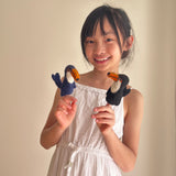 A smiling girl holds two handcrafted needle-felted toucan finger puppets on her fingers against a bright, plain beige background. Made of 100% wool, one toucan has a navy blue body with a white chest and bright orange beak, while the other is black with a white chest and similar orange beak. These unique and playful finger puppets highlight fair-trade craftsmanship from Nepal.