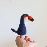 A needle felted toucan finger puppet is playfully held on a finger of a female hand with red polished nails, against a plain white background. This handcrafted toucan features a vibrant orange beak with a black tip, bright blue eyes rimmed with orange, a white neck, and a deep blue body, making it an engaging toy for kids.