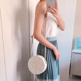 A woman poses for the camera, wearing a white top and light blue skirt, holding her phone in one hand. She wears a round woven rattan crossbody bag on her left shoulder with an adjustable leather strap. The bag features a unique star design and appears to be made of natural rattan materials. It is white in color, round in shape, medium in size, and has a textured appearance.