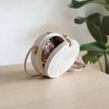 A white rattan bag with a star design and batik lining, featuring solid and patterned sections, has a round shape with textured weaving resembling natural fiber. The bag is opened to showcase its batik lining. A small snap closure with pink leather flap is attached to the top for closing the bag.