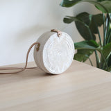 A White Bali Star Round Rattan Crossbody Bag, round-shaped bag made from natural rattan material, featuring a unique star design with handmade details, predominantly white color. The adjustable leather strap is extended on the table, suitable for any occasion.
