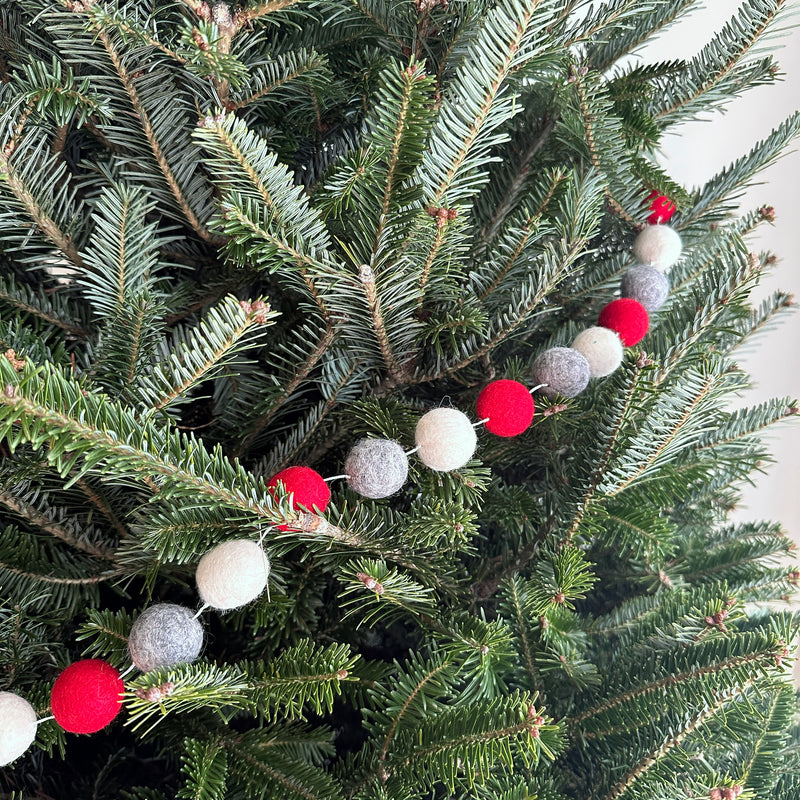 A felt festive pompom ball garland is draped on a lush, green Christmas tree. The garland features alternating red, gray, and white felt balls, adding a warm, celebratory touch to the festive branches.