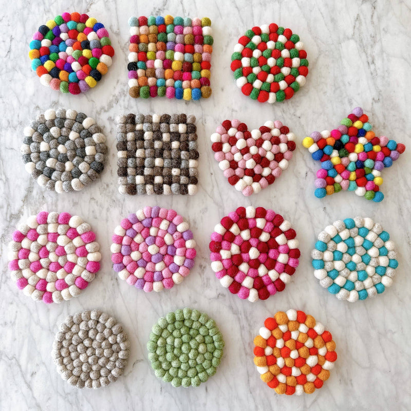 A gray felt ball round placemat is prominently displayed among other felt ball placemats on a bright marble surface. This handcrafted felt placemat, ethically made by Nepalese female artisans, is composed of tightly packed, hand-stitched gray felt balls, each about an inch in size. Other customizable felt pieces in various shapes and colors are also shown, highlighting the use of 100% organic lamb wool and azo-free dyes.