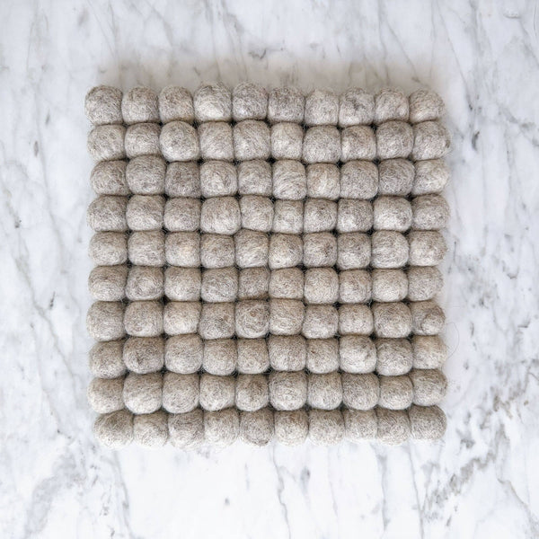 An 8-inch gray felt ball square pot trivet or placemat rests flat on a white marble surface. This handcrafted trivet, made from ethically sourced organic wool felt, features numerous small felt balls meticulously hand-stitched together to form a uniform square shape. Its cozy texture and neutral gray hue provide a calming, natural look, highlighting its fair-trade craftsmanship by Nepalese female artisans.