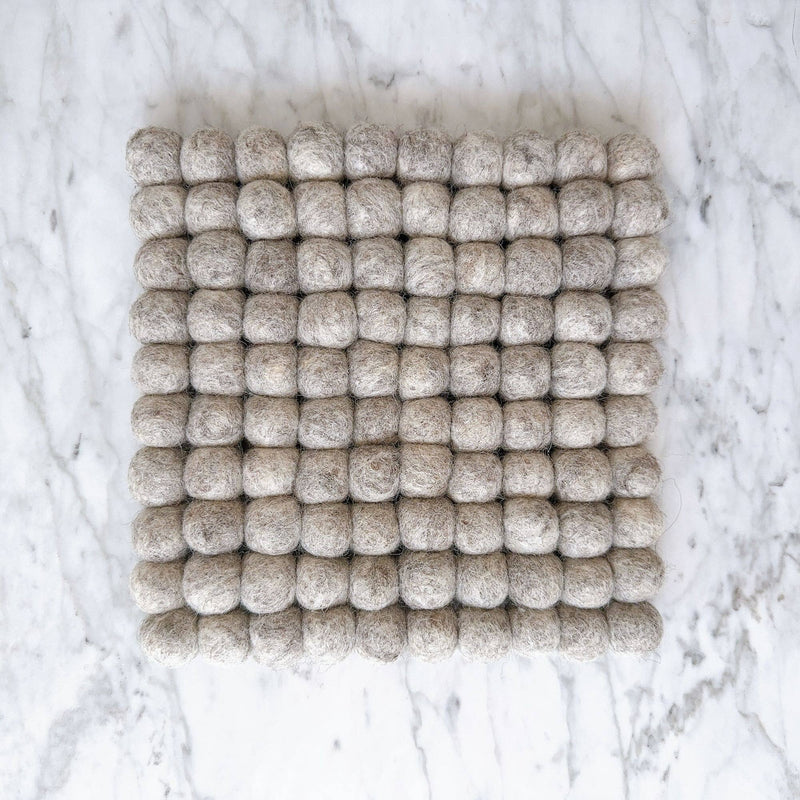 An 8-inch gray felt ball square pot trivet or placemat rests flat on a white marble surface. This handcrafted trivet, made from ethically sourced organic wool felt, features numerous small felt balls meticulously hand-stitched together to form a uniform square shape. Its cozy texture and neutral gray hue provide a calming, natural look, highlighting its fair-trade craftsmanship by Nepalese female artisans.
