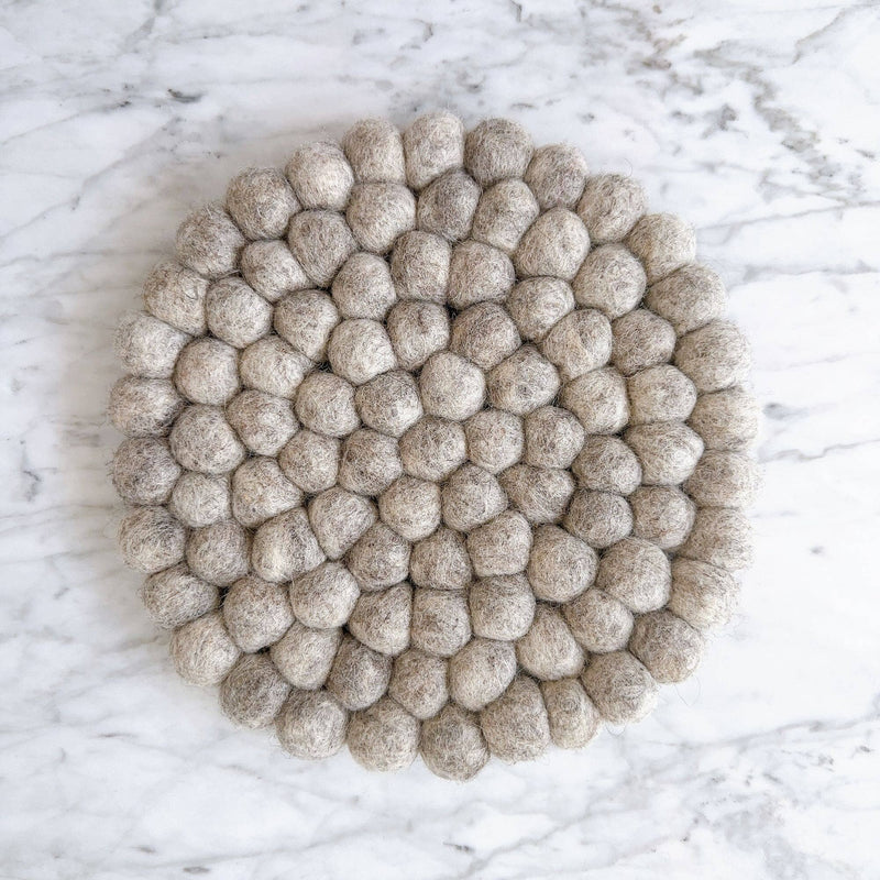 A round gray felt ball placemat, doubling as a pot trivet, rests on a clean white marble surface. It is densely packed with hand-stitched organic wool felt balls, creating a textured surface with subtle variations in gray tones. Ethically handmade by Nepalese female artisans, this handcrafted felt placemat adds a cozy, rustic elegance to home decor.