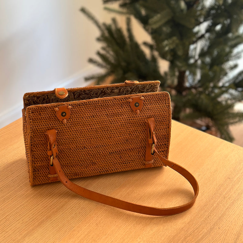 A woven rattan handbag, Bordeaux color, resting on a wooden table amidst a cozy Christmas setting. The bag features two adjustable leather handles, a snap closure, and a rectangular shape with rounded corners.
