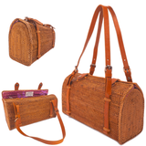 A beautifully crafted Bali rattan handbag, presented on a neutral background, features a woven brown color with visible stitching details. The bag boasts a prominent adjustable leather handle, a rectangular shape with rounded corners, and a slightly tapered body. The interior lining is a solid color, possibly matching the exterior's natural tones.