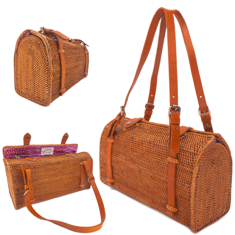A beautifully crafted Bali rattan handbag, presented on a neutral background, features a woven brown color with visible stitching details. The bag boasts a prominent adjustable leather handle, a rectangular shape with rounded corners, and a slightly tapered body. The interior lining is a solid color, possibly matching the exterior's natural tones.