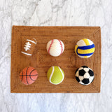 Six vibrant handmade felt ball ornaments, including a tennis ornament, a basketball ornament, a volleyball ornament, a soccer ornament, and a baseball ornament, rest on a woven brown placemat, set against a clean white marble background. This wool felt ball ornament set features classic lines, accurately depicting each ball. Each one of them includes a jute string for hanging, making it a cheerful Christmas decoration.