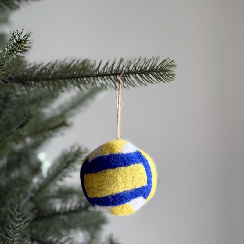 A spherical wool felt Volleyball ornament from the American Popular Sports Ornament Set hangs on a blurred Christmas tree. It features alternating black and white pentagons, crafted from soft, textured 100% wool felt. A jute string is attached for graceful hanging.