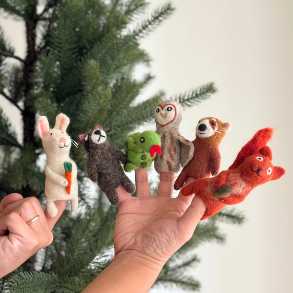 A hand displays a set of six assorted forest animal felt finger puppets against a bright background with a Christmas pine tree. This handmade felt finger puppet collection includes a white bunny holding a carrot, a dark brown raccoon, a green snake with a red tongue, a tan and brown owl, a light brown red panda, and a red squirrel with a bushy tail. These unique finger puppets are perfect for imaginative play and storytelling.
