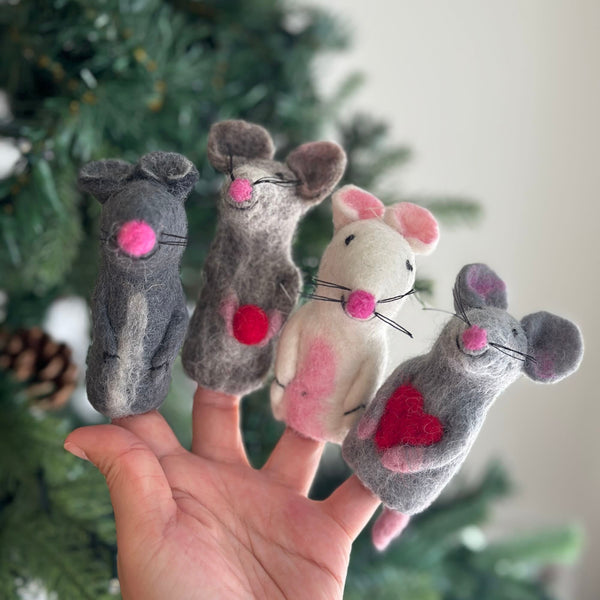 A set of four assorted wool felt mice finger puppets is presented by a female hand, set against a blurred, festive Christmas tree background. These unique handcrafted needle felted mice showcase a variety of colors: solid gray, gradient gray holding a red felt ball, white with pink ears and a chest spot, and gray holding a red heart. Each charming mouse features a pink nose and black whiskers, highlighting the intricate detail of these fair-trade toys.