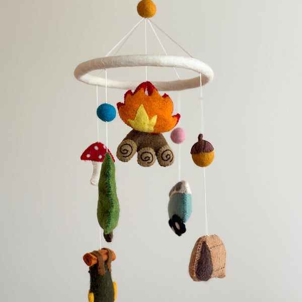 A vibrant camping-themed wool felt baby mobile is displayed against a plain white background. Handcrafted figures made from natural New Zealand sheep wool and eco-friendly dyes hang from a white felt ring topped with a yellow felt ball. Suspended at various lengths are a cozy campfire with orange and yellow flames, a blue minivan, a tall green tree, a brown and tan tent, a dark green backpack, a bear-adorned kayak, and blue and pink balls.