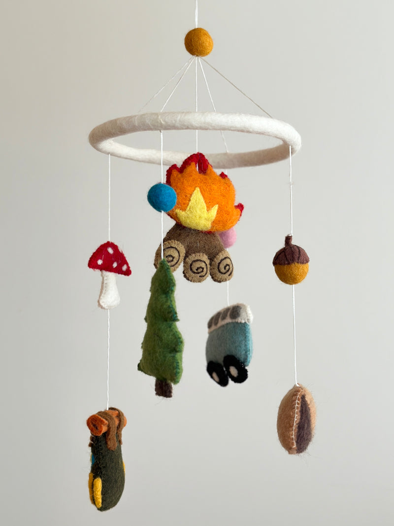 A handcrafted wool felt camping baby mobile is displayed, featuring a white felt ring suspending various camping-themed figures. Crafted from natural New Zealand sheep wool, these include a cozy campfire with orange and yellow flames, a green pine tree, a charming blue minivan with black wheels, and a green, brown, and yellow hiking backpack. A yellow felt ball tops the mobile, completing the outdoor adventure theme.