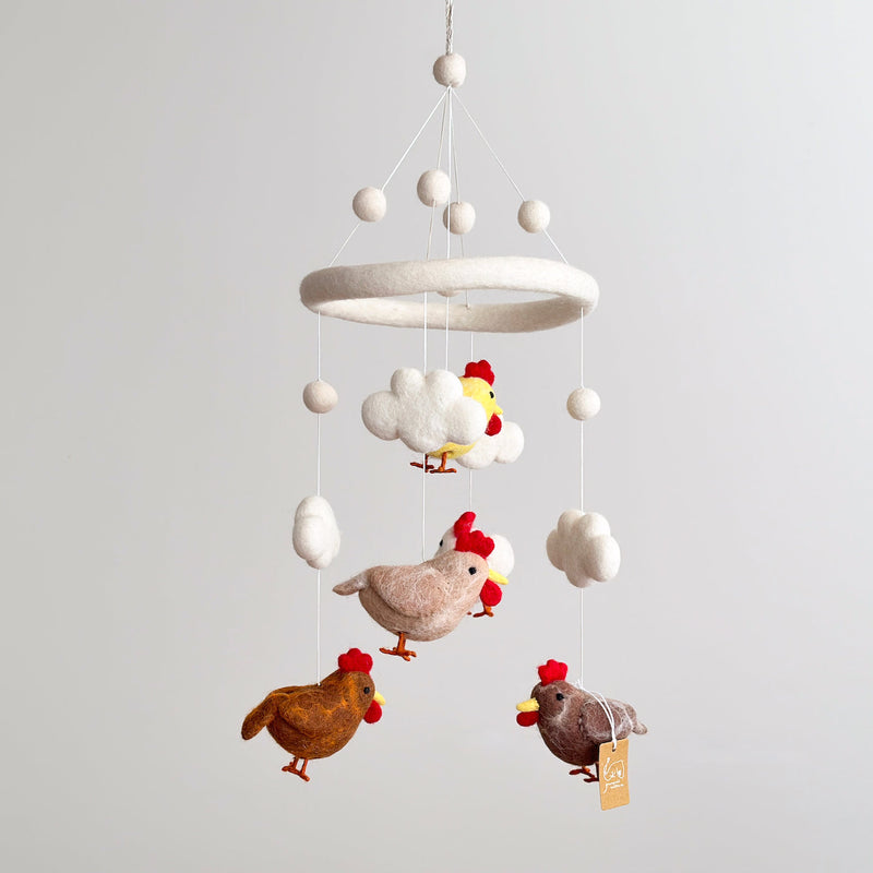 A baby mobile, made from wool felt, hangs from the ceiling against a neutral background. The mobile features five colorful chickens with different patterns: brown, white, and yellow. Each chicken is attached by a string and appears to float in mid-air, surrounded by small clouds made from soft material.