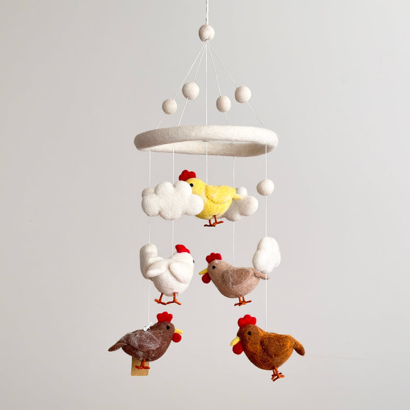 A baby mobile featuring five colorful chickens made from wool felt, each suspended on a string, hangs from the ceiling. The chickens are depicted in a cartoon-like style with one wearing a red hat. A small cloud-shaped element is visible below the chickens.