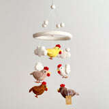 A baby mobile featuring five colorful chickens, each with a different pose and color, hangs from the ceiling. The soft, felt-like material of the chickens has a plush texture. A minimalist white wall provides a neutral backdrop for the vibrant colors of the mobile.