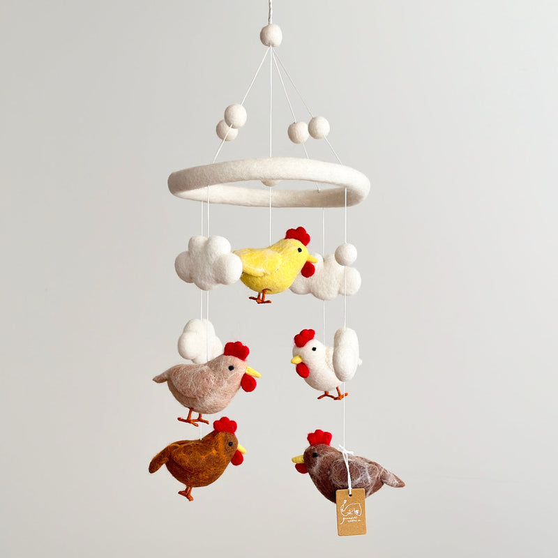 A baby mobile featuring five colorful chickens, each with a different pose and color, hangs from the ceiling. The soft, felt-like material of the chickens has a plush texture. A minimalist white wall provides a neutral backdrop for the vibrant colors of the mobile.
