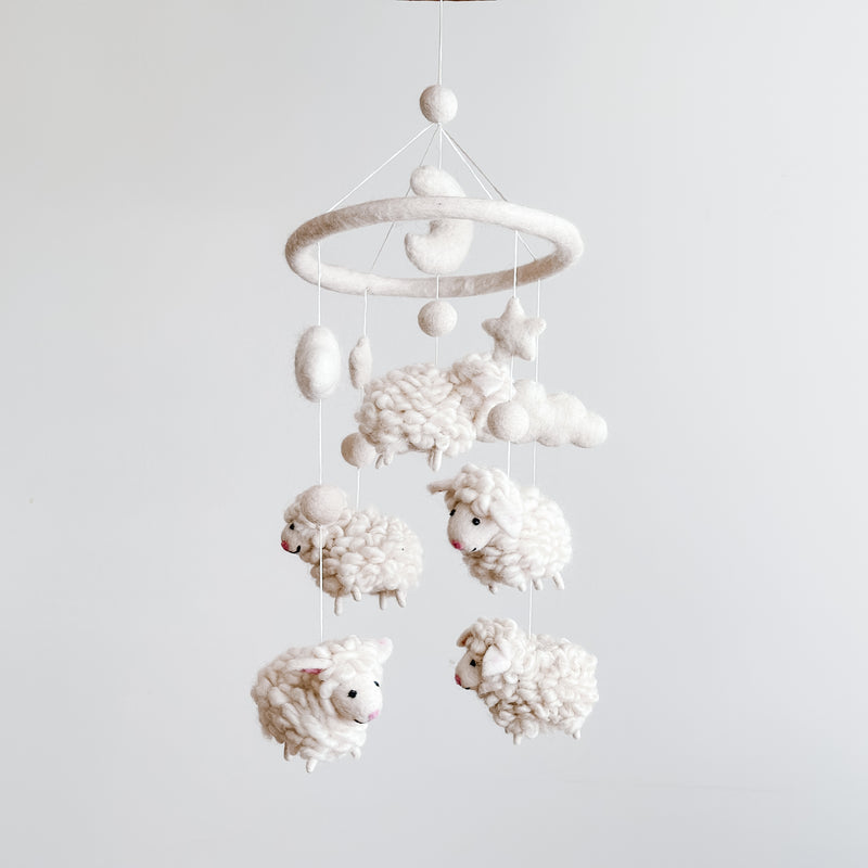A Counting Sheep wool felt baby mobile is suspended against a plain white background. It features a circular felt hoop from which several fluffy white sheep hang by delicate white threads. Each soft, organic wool felt sheep has small, cute faces, adding a gentle and playful touch for a baby's nursery.