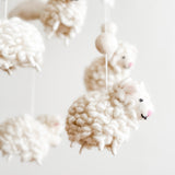 A close-up of a Counting Sheep wool felt baby mobile hangs against a bright, neutral background, emphasizing its gentle aesthetic. The mobile features a felt hoop from which several fluffy, white wool felt sheep are suspended by thin white strings. Each sheep has a rounded, textured body with small pink details for its nose and inner ears, separated by white felt balls.