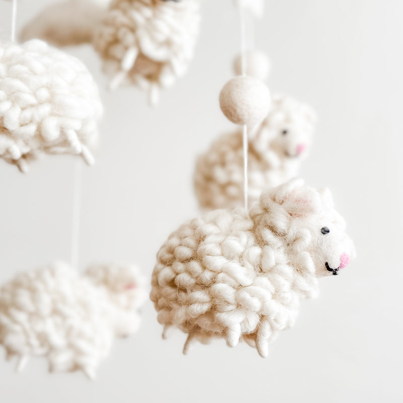 A close-up of a Counting Sheep wool felt baby mobile hangs against a bright, neutral background, emphasizing its gentle aesthetic. The mobile features a felt hoop from which several fluffy, white wool felt sheep are suspended by thin white strings. Each sheep has a rounded, textured body with small pink details for its nose and inner ears, separated by white felt balls.