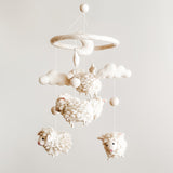 A soothing Counting Sheep wool felt baby mobile is displayed against a light gray background, showcasing its soft design. Fluffy white wool felt sheep are suspended at varying heights from a circular felt hoop, creating a calming nursery aesthetic.