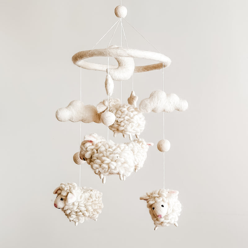 A soothing Counting Sheep wool felt baby mobile is displayed against a light gray background, showcasing its soft design. Fluffy white wool felt sheep are suspended at varying heights from a circular felt hoop, creating a calming nursery aesthetic.