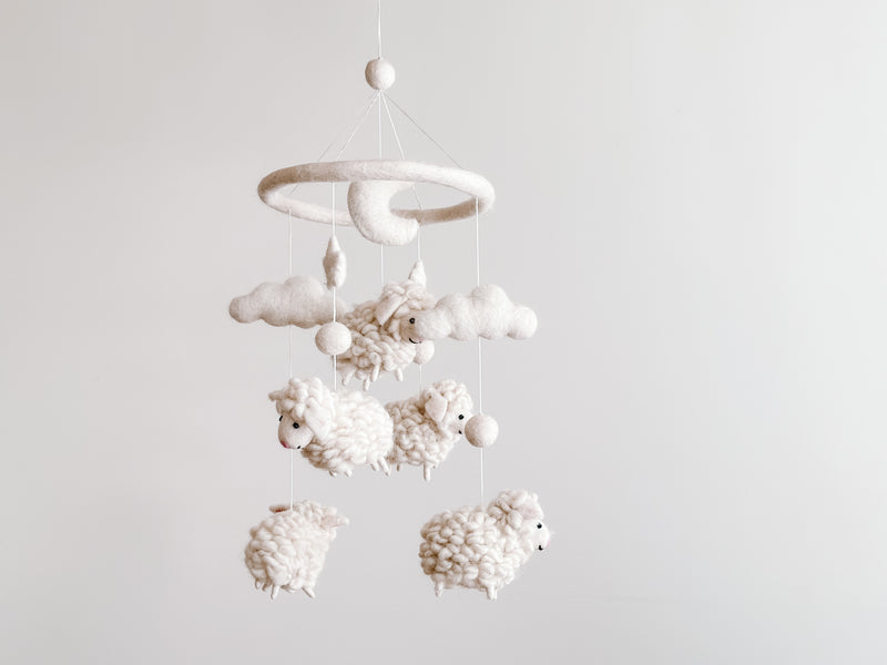 A handcrafted Counting Sheep wool felt baby mobile hangs against a plain white backdrop, creating a calm atmosphere. The mobile features a circular white felt hoop from which several fluffy white sheep figures are suspended by thin white strings. Each sheep is crafted from soft, organic wool felt, emphasizing its wool-like texture.