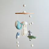 A colorful wool felt baby mobile, featuring a white jellyfish and various sea creatures, hangs against a plain light gray wall. Suspended from a wooden frame by white threads are teal whales, a white jellyfish with dotted details, a gray seahorse, and a green sea turtle. White wool felt clouds and balls accent the top and bottom of the mobile.