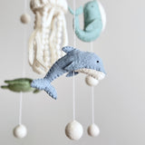 A wool felt baby mobile, featuring a circular wooden frame, is centered against a light gray background. Suspended by thin cotton threads are various wool felt sea creatures, including a blue dolphin with a white underbelly, a white jellyfish with dangling tentacles, a light blue whale, and a green sea turtle. White felt balls hang below each soft, textured creature, creating a calm nursery baby mobile.