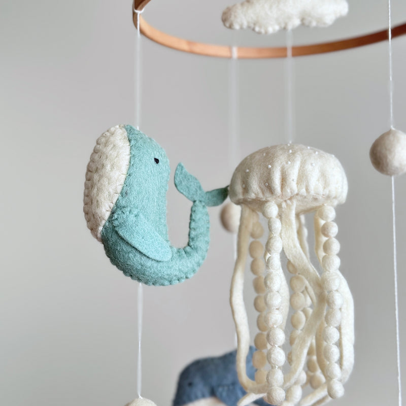 A jellyfish wool felt baby mobile is prominently displayed in a close-up, featuring a white jellyfish and a teal whale suspended from a wooden frame against a light gray background. The white jellyfish has a rounded top adorned with small white beads and long, dangling felt ball tentacles. Next to it, a charming teal felt whale with a white belly and a small tail adds to the serene, handmade quality, perfect for a baby's nursery.