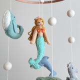 A felt baby mobile, featuring a mermaid and various sea creatures, hangs from a string, suspended in mid-air. The mobile is made of wool felt and has a serene, tranquil atmosphere, evoking the beauty of marine life. The product's neutral background allows the mobile to be the focal point, suggesting it would be used to decorate a nursery or child's room.